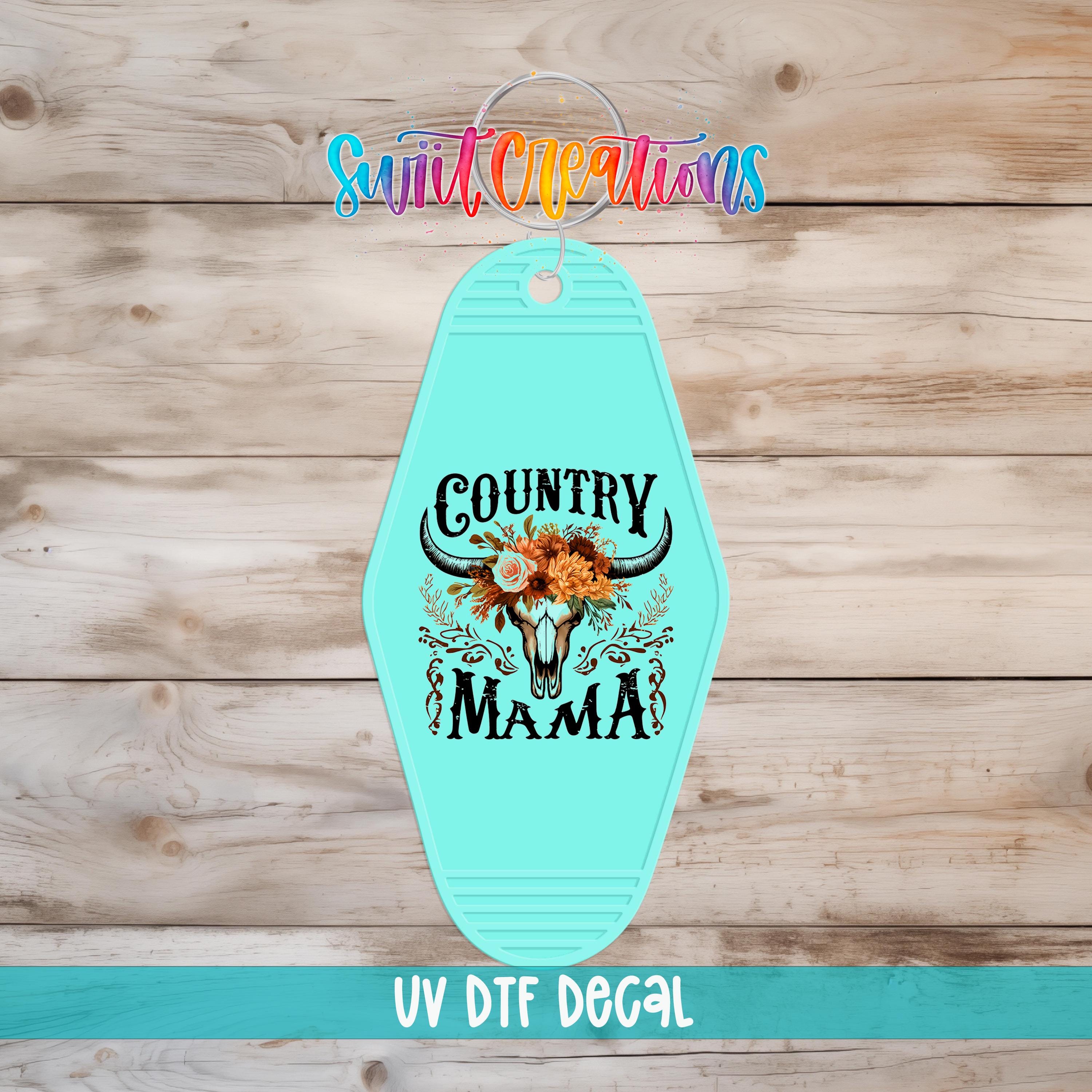A turquoise keychain with a decorative design featuring a cow's head and the text "COUNTRY MAMA" printed on it, placed on a wooden surface.