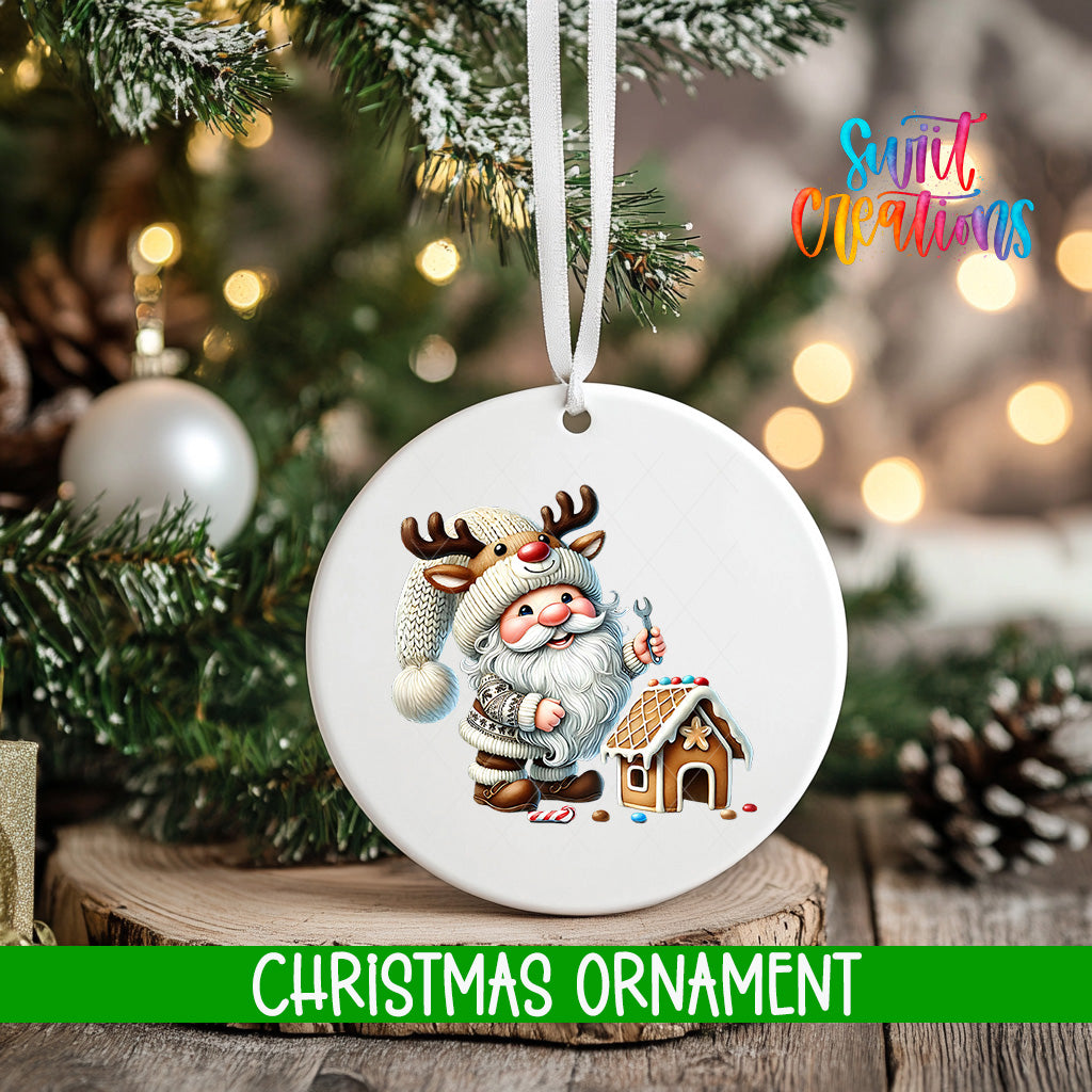 A white Christmas ornament featuring a Santa Claus figure with antlers and a gingerbread house, hanging on a tree branch with a blurred background of lights.