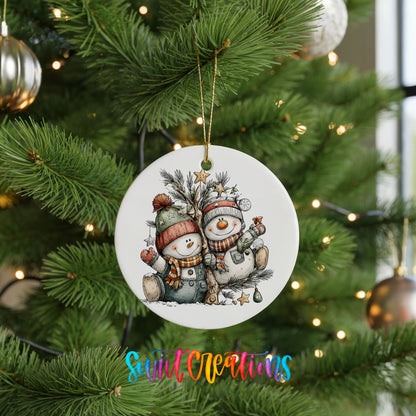 A white Christmas tree ornament featuring two snowmen, one wearing a green hat and the other a red hat, is hanging from the tree.