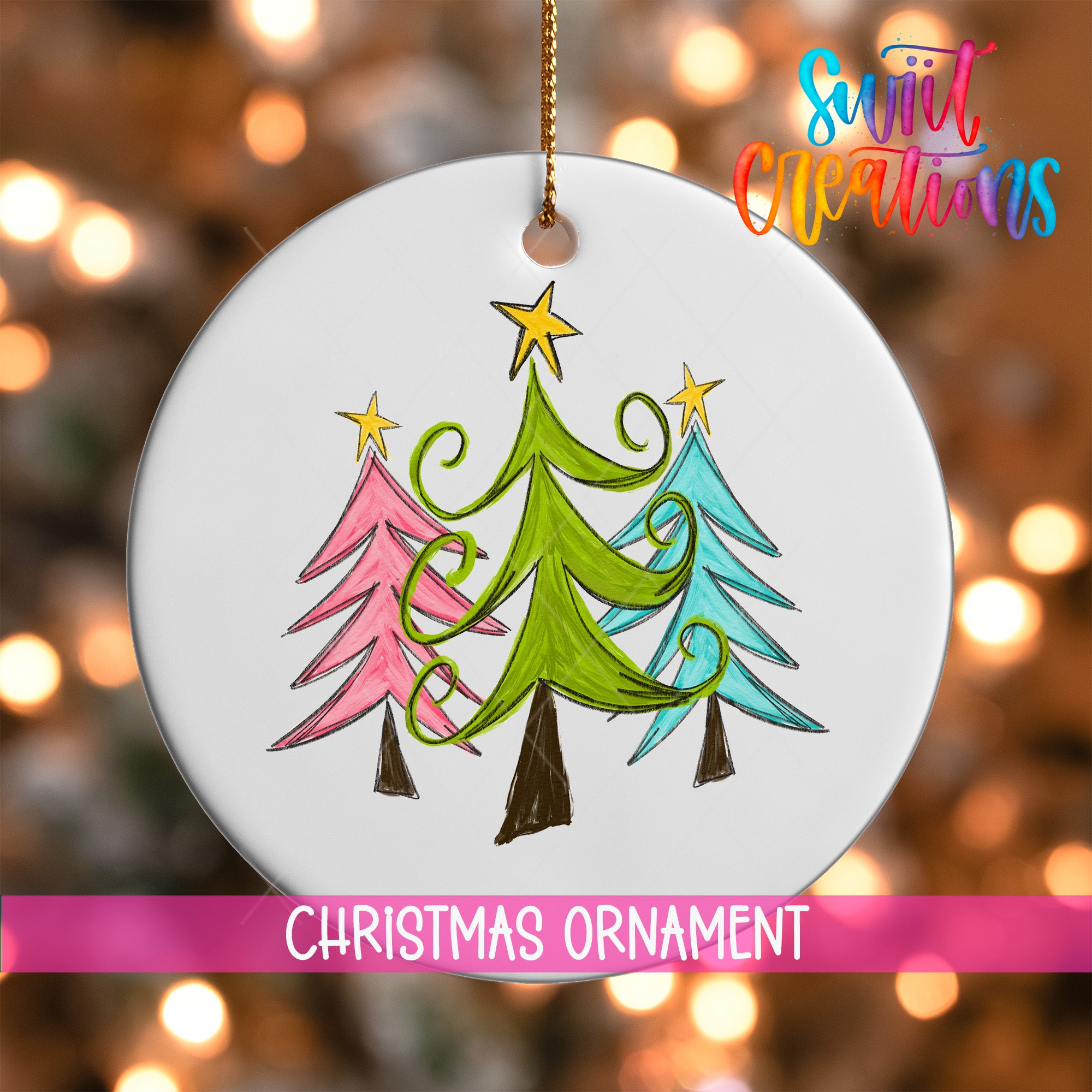 A white Christmas ornament with a drawing of three stylized Christmas trees in different colors, including pink, green, and blue, hanging against a blurred background with colorful lights.