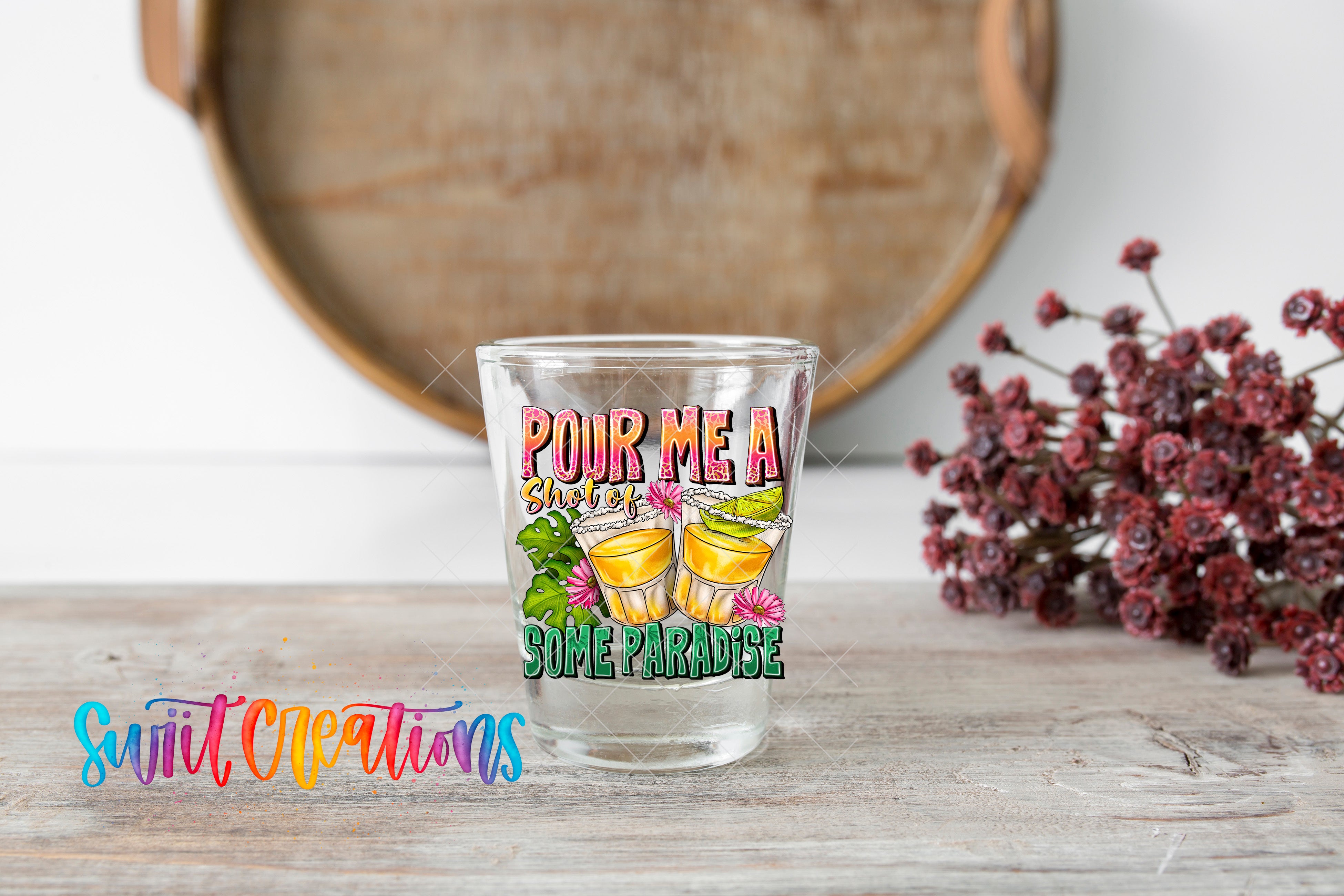 A shot glass with a colorful design and text, placed on a wooden surface next to a bunch of red flowers.