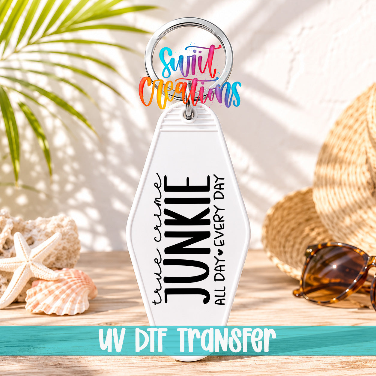 A keychain with the text "True Crime Junkie All Day Every Day" printed on it, along with a colorful ring and a straw hat. The keychain is placed on a wooden surface, with sunglasses and a palm frond visible in the background.
