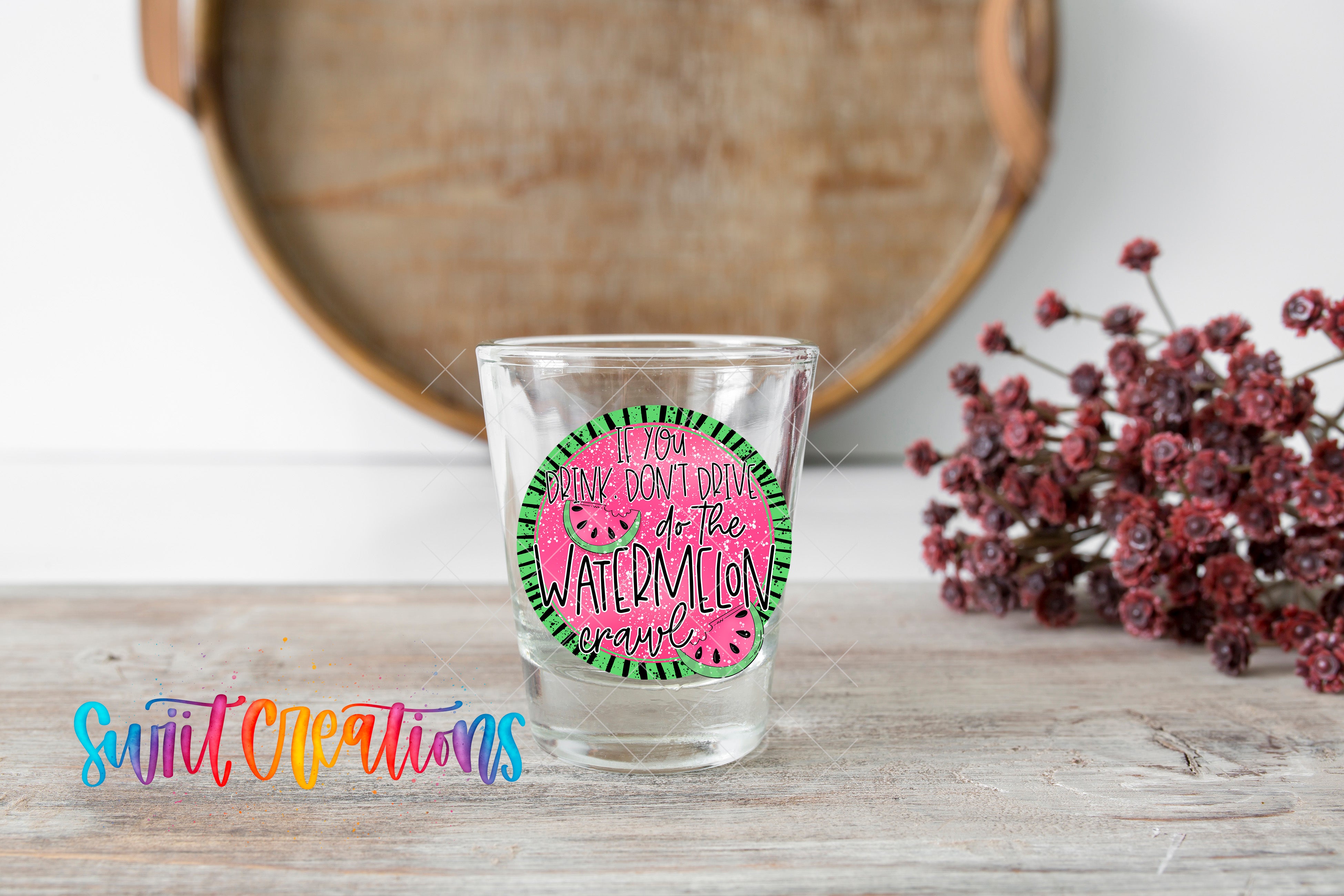 A shot glass with a watermelon sticker on it, placed on a wooden surface next to a bunch of red flowers.