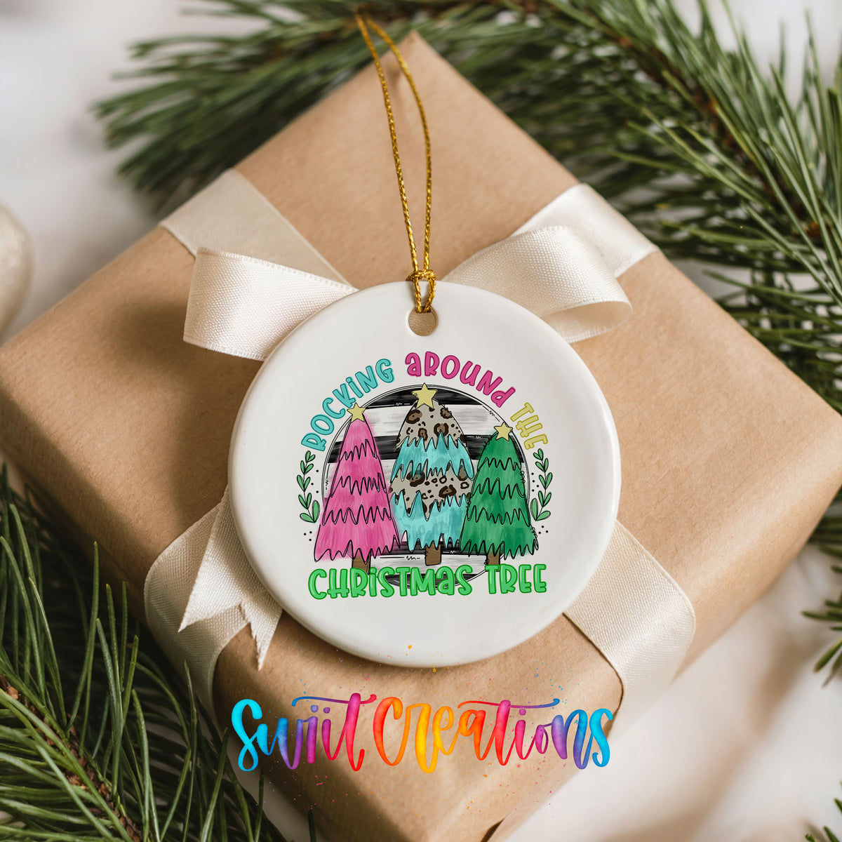 A white ceramic ornament with a colorful design hangs from a ribbon, surrounded by pine branches.