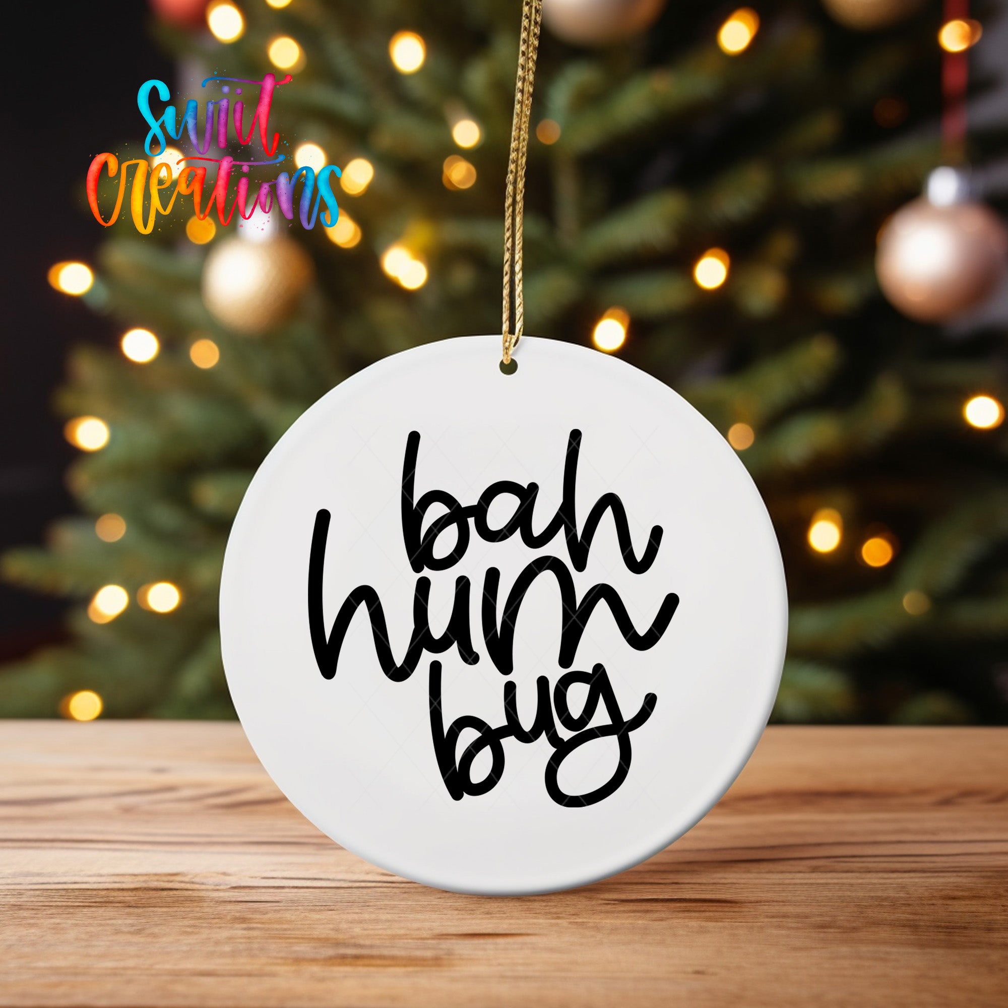 A white ornament with the text "bah hum bug" hanging from a string, placed in front of a blurred Christmas tree with colorful lights.