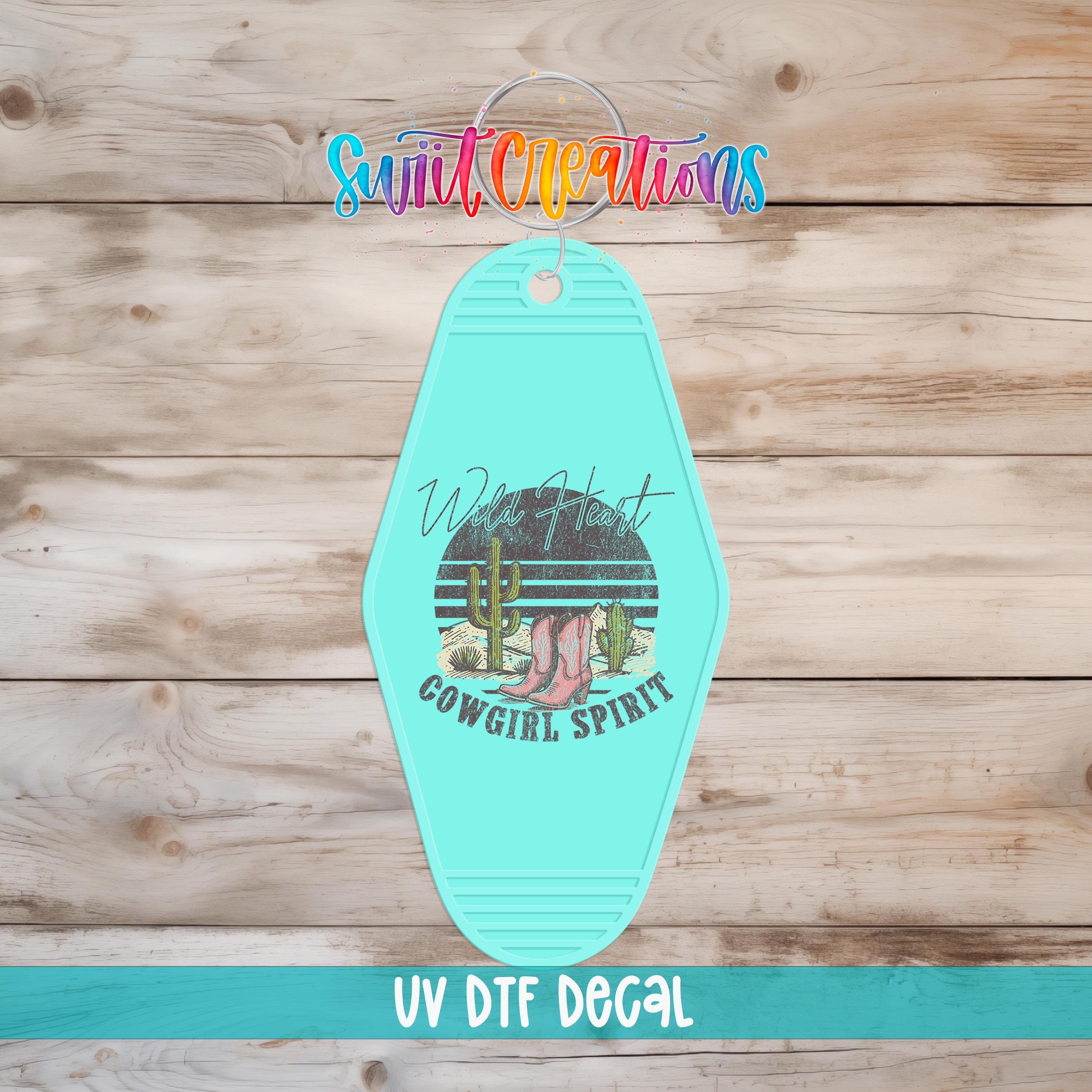 A turquoise keychain with a graphic design featuring a cactus and the text "Wild Heart Cowgirl Spirit" is displayed against a wooden background.