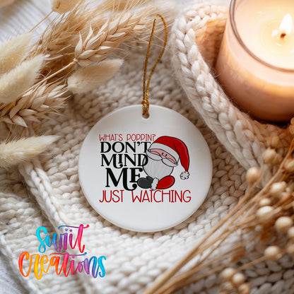 A white ornament with a Santa Claus design and text that says "What's Popping? Don't Mind Me Just Watching" is displayed on a knitted surface next to a lit candle.