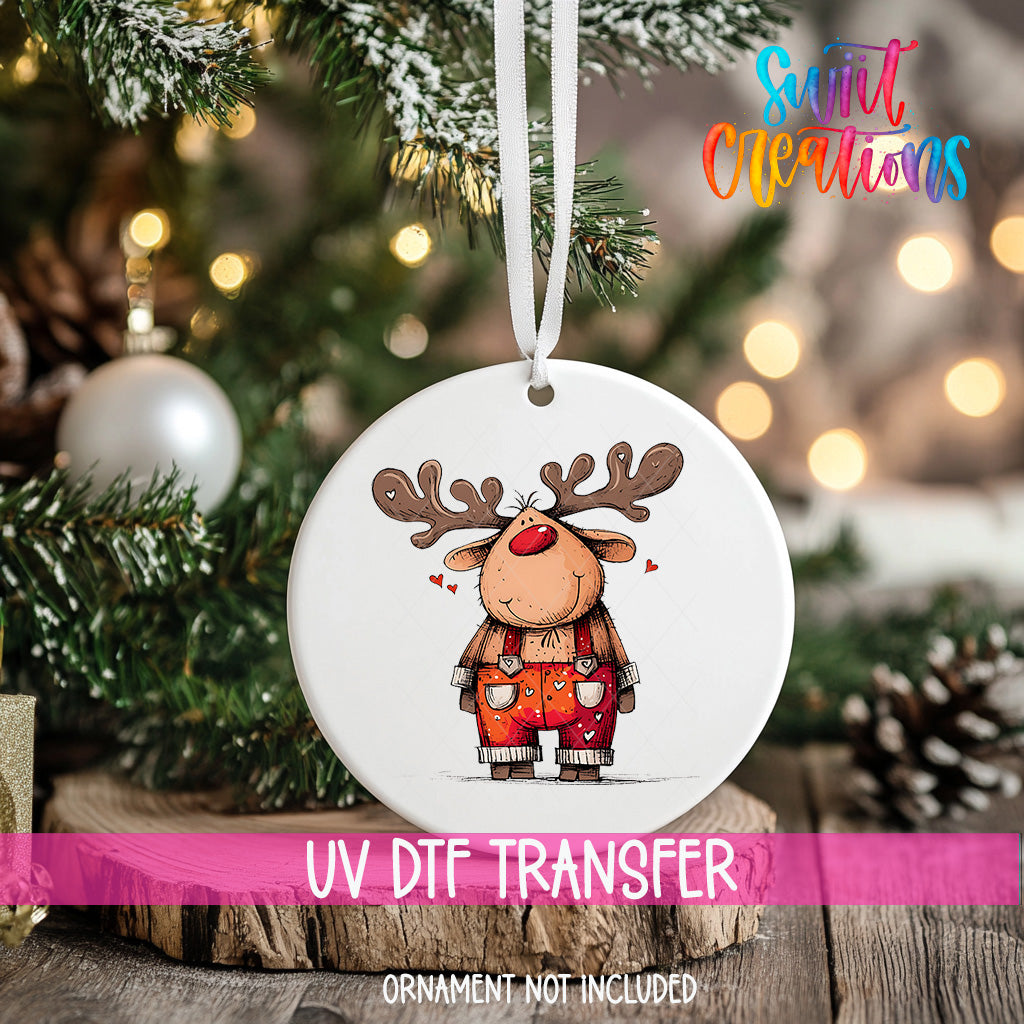 A white ornament with a cartoon reindeer design hangs on a Christmas tree, surrounded by pine branches and twinkling lights.