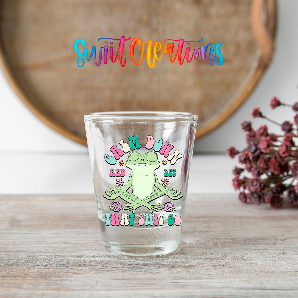 Calm Down And Let That Sh*t Go shot glass transfer on a clear glass beside a wooden barrel and dried flowers