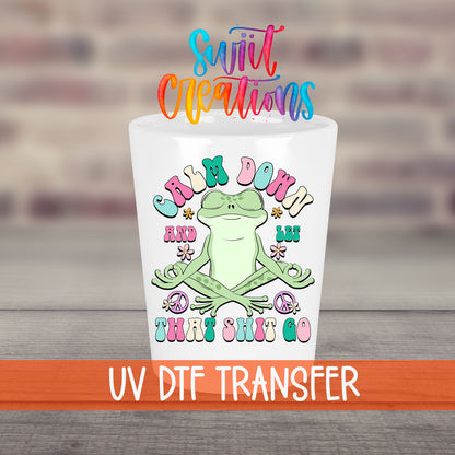 Calm Down And Let That Sh*t Go UV DTF transfer on a white shot glass in front of a brick wall