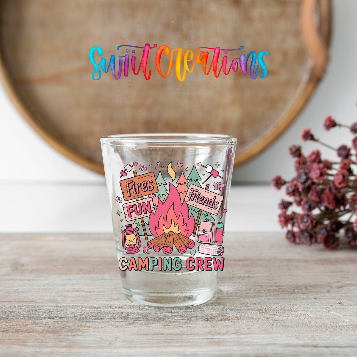 Camping Crew shot glass transfer on a clear glass beside a wooden barrel and dried flowers