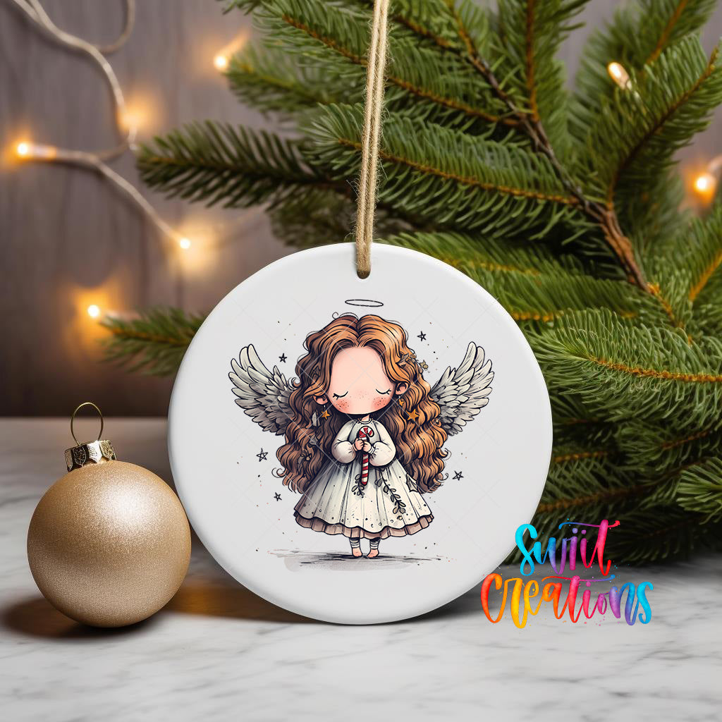 A white ceramic ornament featuring a cartoon illustration of a young girl with curly hair and wings, holding a halo and a staff, is displayed on a table next to a gold ornament and a Christmas tree.
