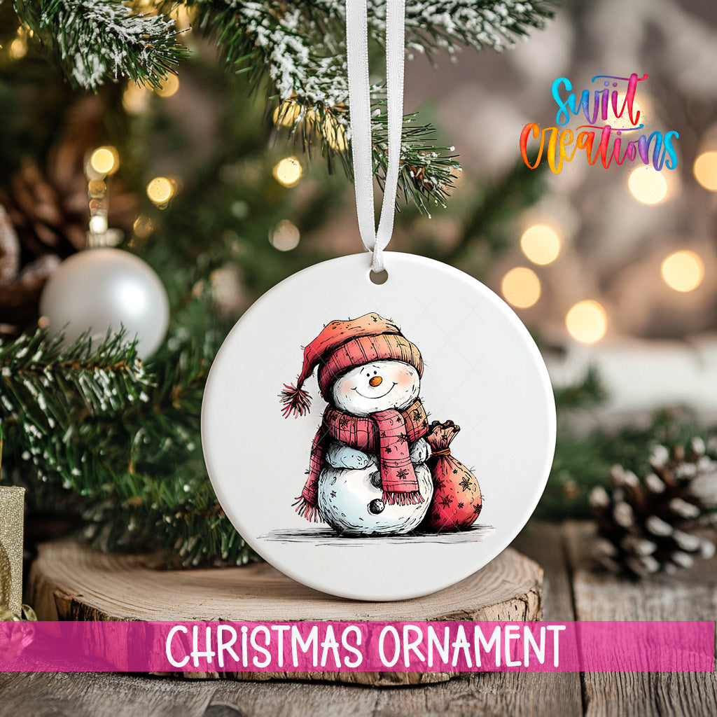 A white Christmas ornament featuring a snowman is hanging from a tree branch, with a blurred background of lights and greenery.