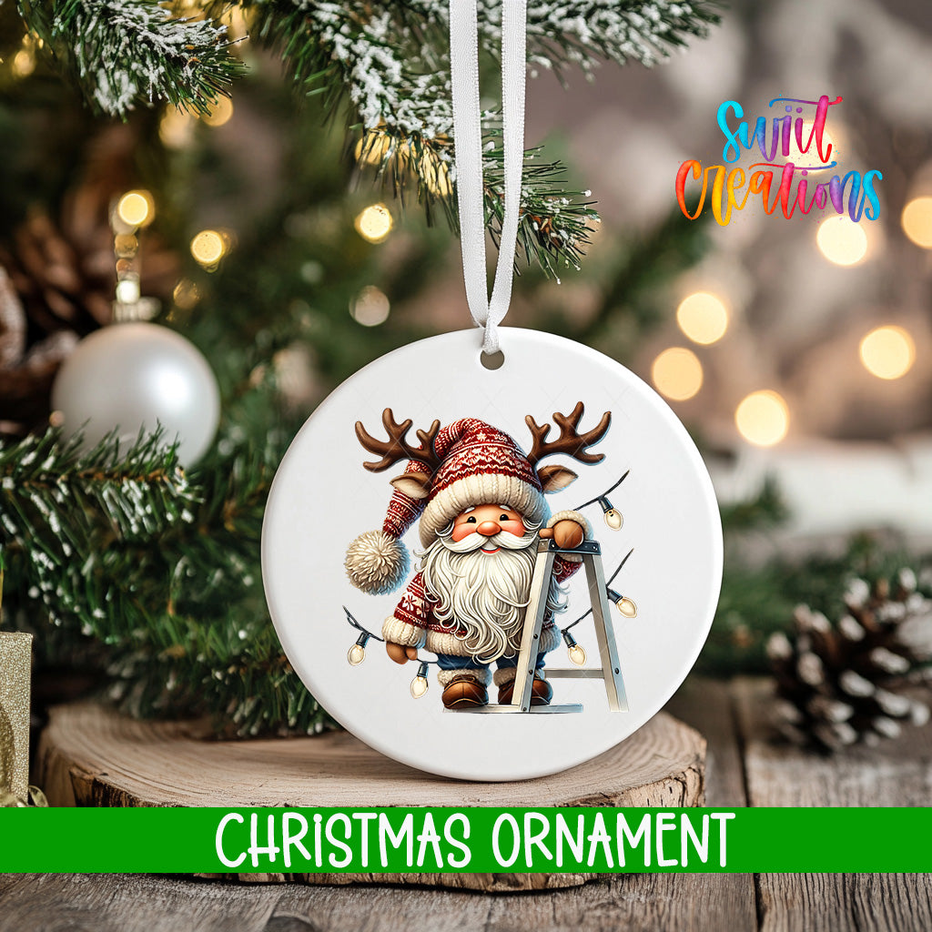 A white Christmas ornament featuring a smiling Santa Claus with antlers and a ladder, hanging on a tree branch with a blurred background of lights.