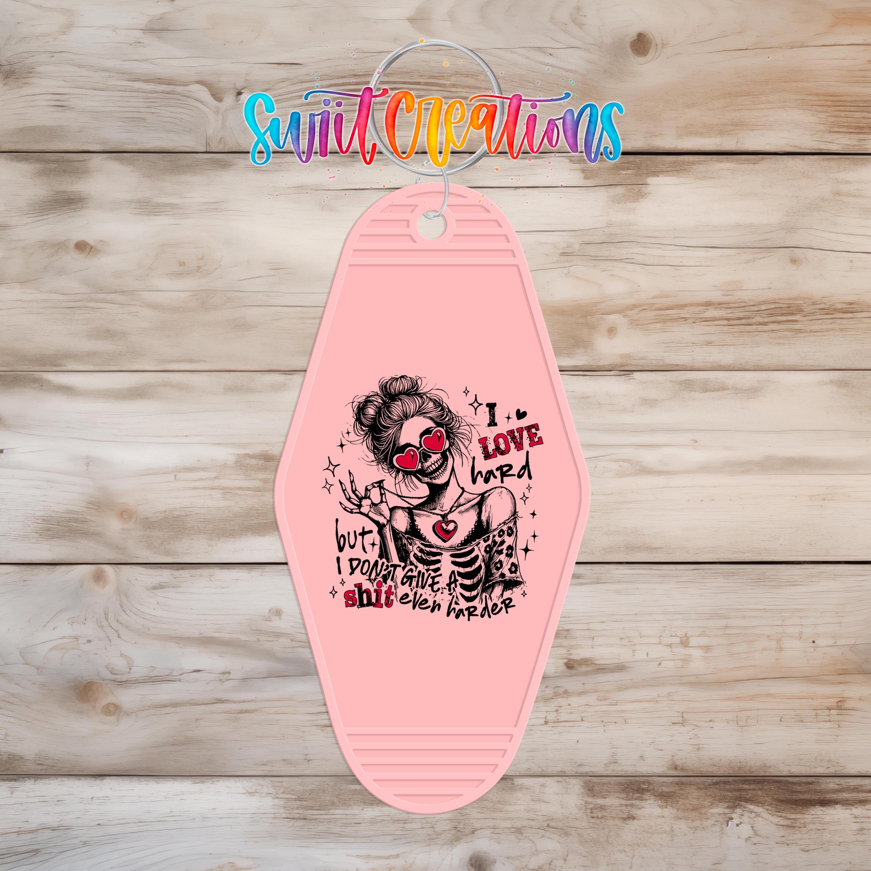 A pink keychain with a graphic design featuring a woman with a skull-like face and the text "I LOVE HARD HARDER" printed on it.