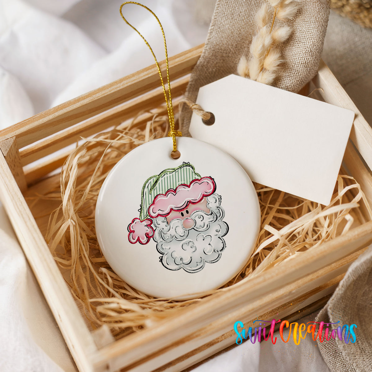 A white ceramic ornament with a Santa Claus face design is displayed in a wooden crate, accompanied by a white tag.