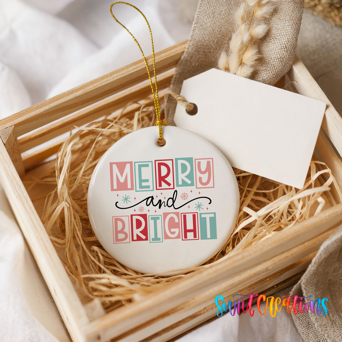 Round ceramic Christmas ornament featuring pastel block-letter Merry and Bright holiday typography, presented in a rustic wooden gift crate