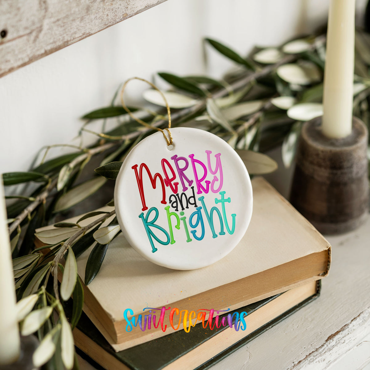 Round ceramic Christmas ornament featuring colorful hand-lettered Merry and Bright holiday typography, styled on books with holiday greenery