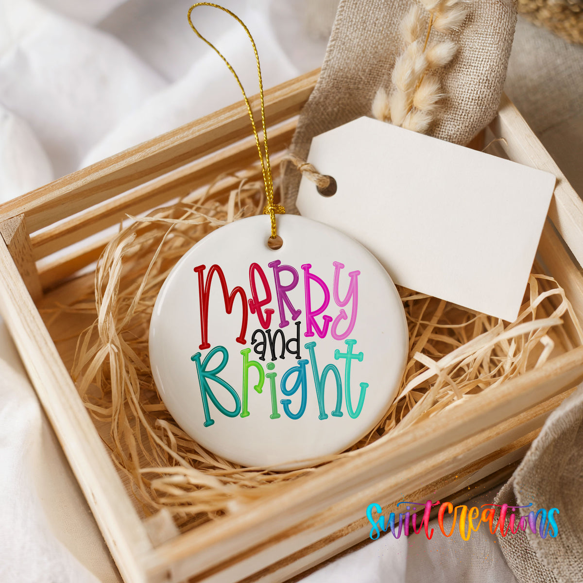 Round ceramic Christmas ornament featuring colorful hand-lettered Merry and Bright holiday typography, presented in a rustic wooden gift crate