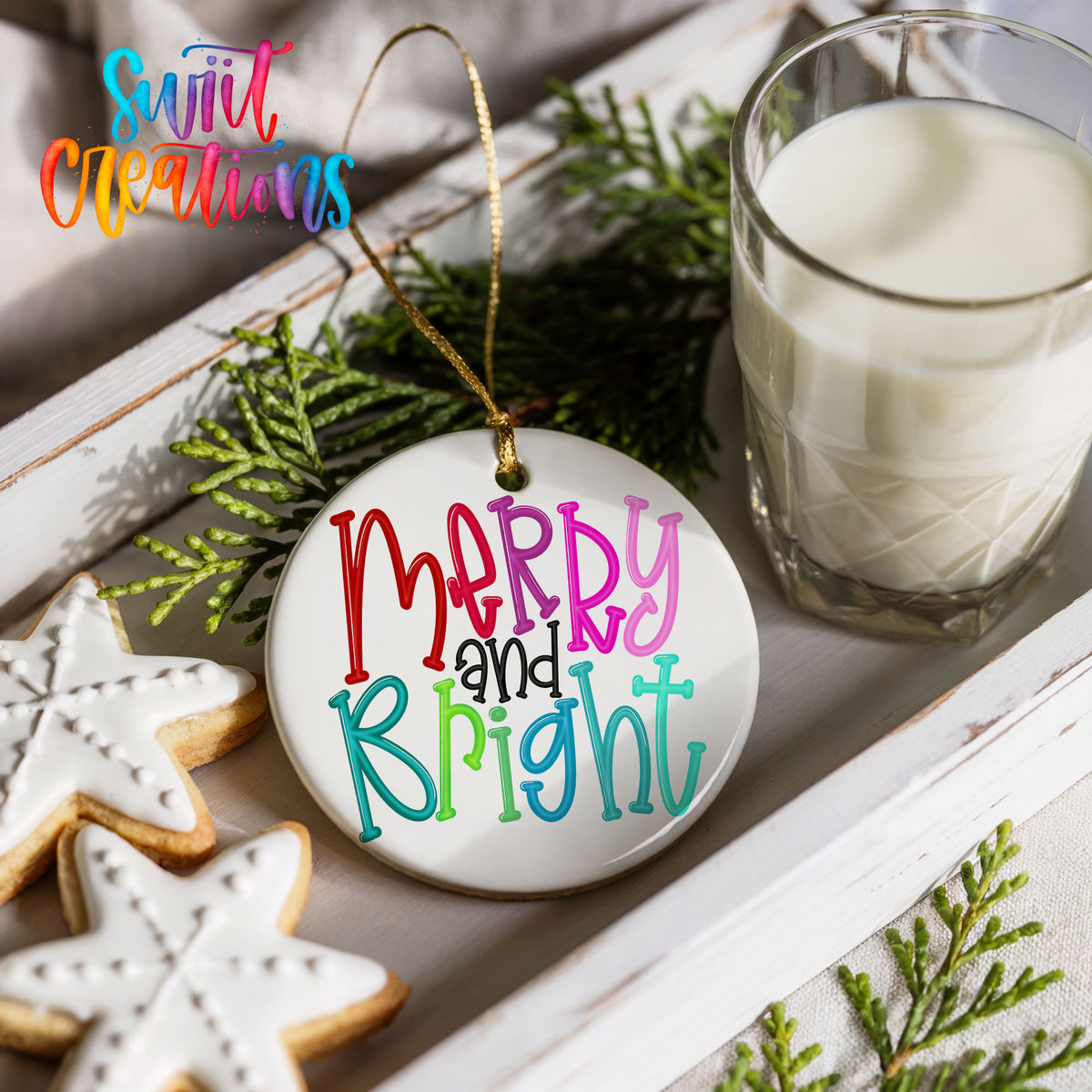 Round ceramic Christmas ornament featuring colorful hand-lettered Merry and Bright holiday typography, styled with cookies and milk
