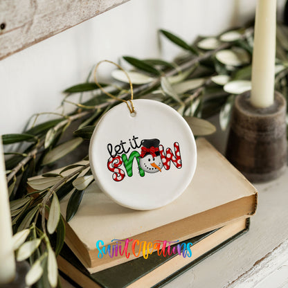 Round ceramic Christmas ornament featuring playful red and black Let It Snow holiday typography, styled on books with holiday greenery