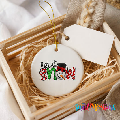 Round ceramic Christmas ornament featuring playful red and black Let It Snow holiday typography, presented in a rustic wooden gift crate
