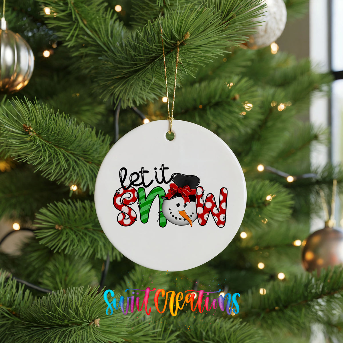Round ceramic Christmas ornament featuring playful red and black Let It Snow holiday typography, hanging on a green Christmas tree