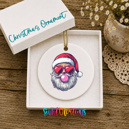 Round ceramic Christmas ornament featuring retro Santa wearing sunglasses and blowing pink bubblegum, displayed in the included gift box