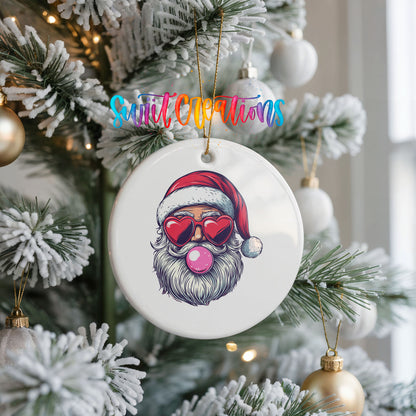 Round ceramic Christmas ornament featuring retro Santa wearing sunglasses and blowing pink bubblegum, hanging on a frosted Christmas tree