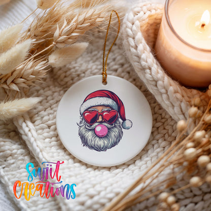 Round ceramic Christmas ornament featuring retro Santa wearing sunglasses and blowing pink bubblegum, styled beside a candle and cozy knit blanket