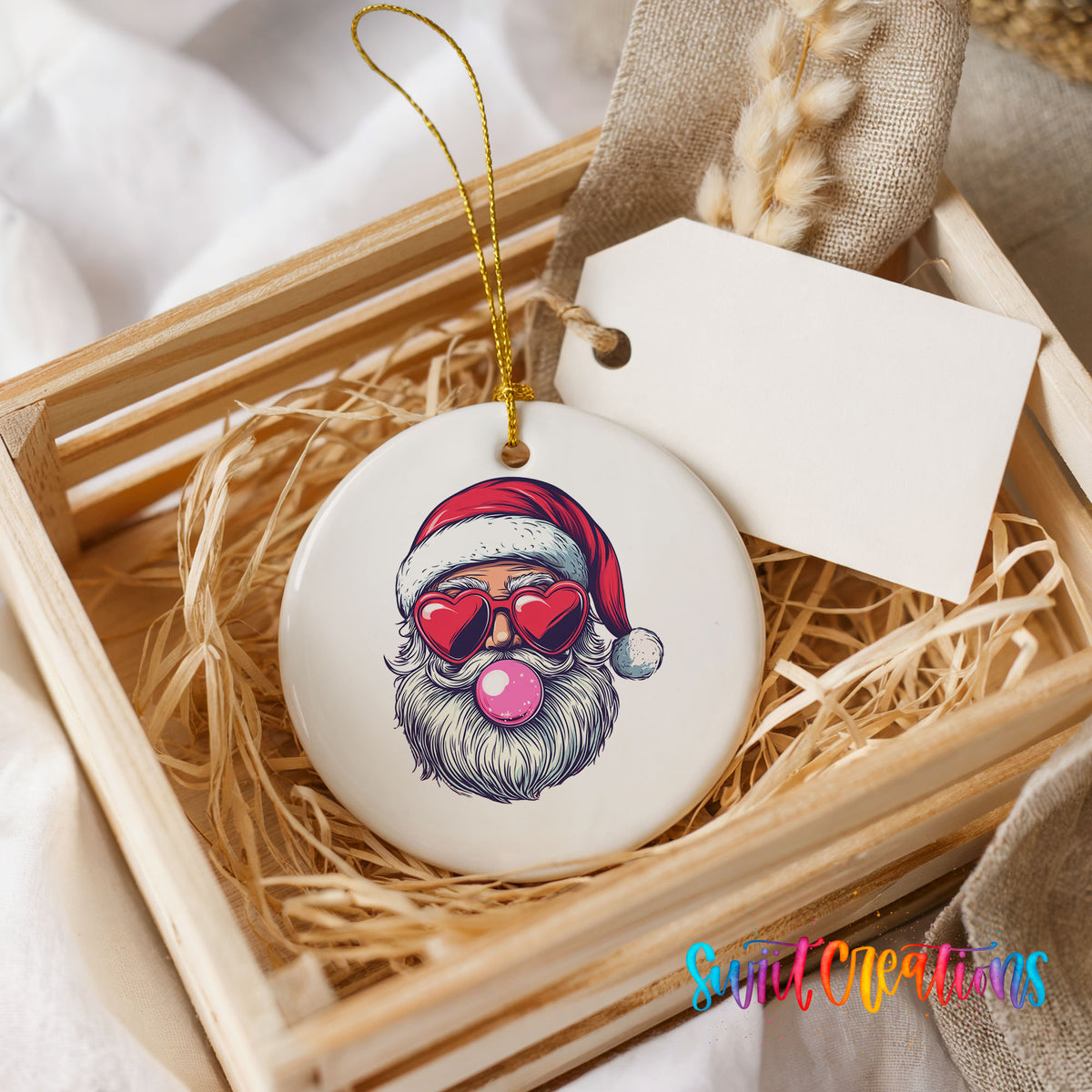Round ceramic Christmas ornament featuring retro Santa wearing sunglasses and blowing pink bubblegum, presented in a rustic wooden gift crate