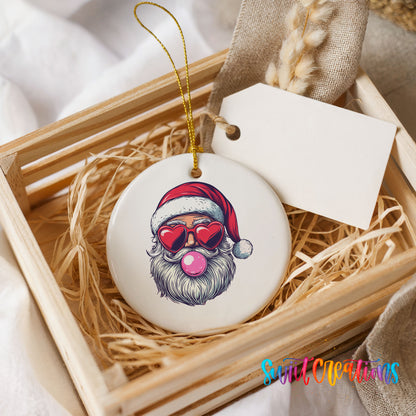 Round ceramic Christmas ornament featuring retro Santa wearing sunglasses and blowing pink bubblegum, presented in a rustic wooden gift crate