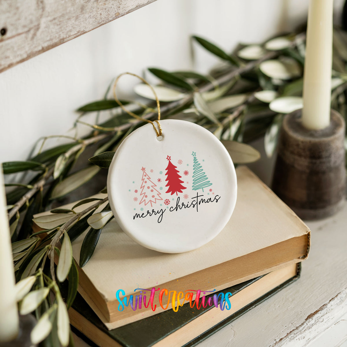 Round ceramic Christmas ornament featuring minimalist red and mint Christmas trees with Merry Christmas script, styled on books with holiday greenery