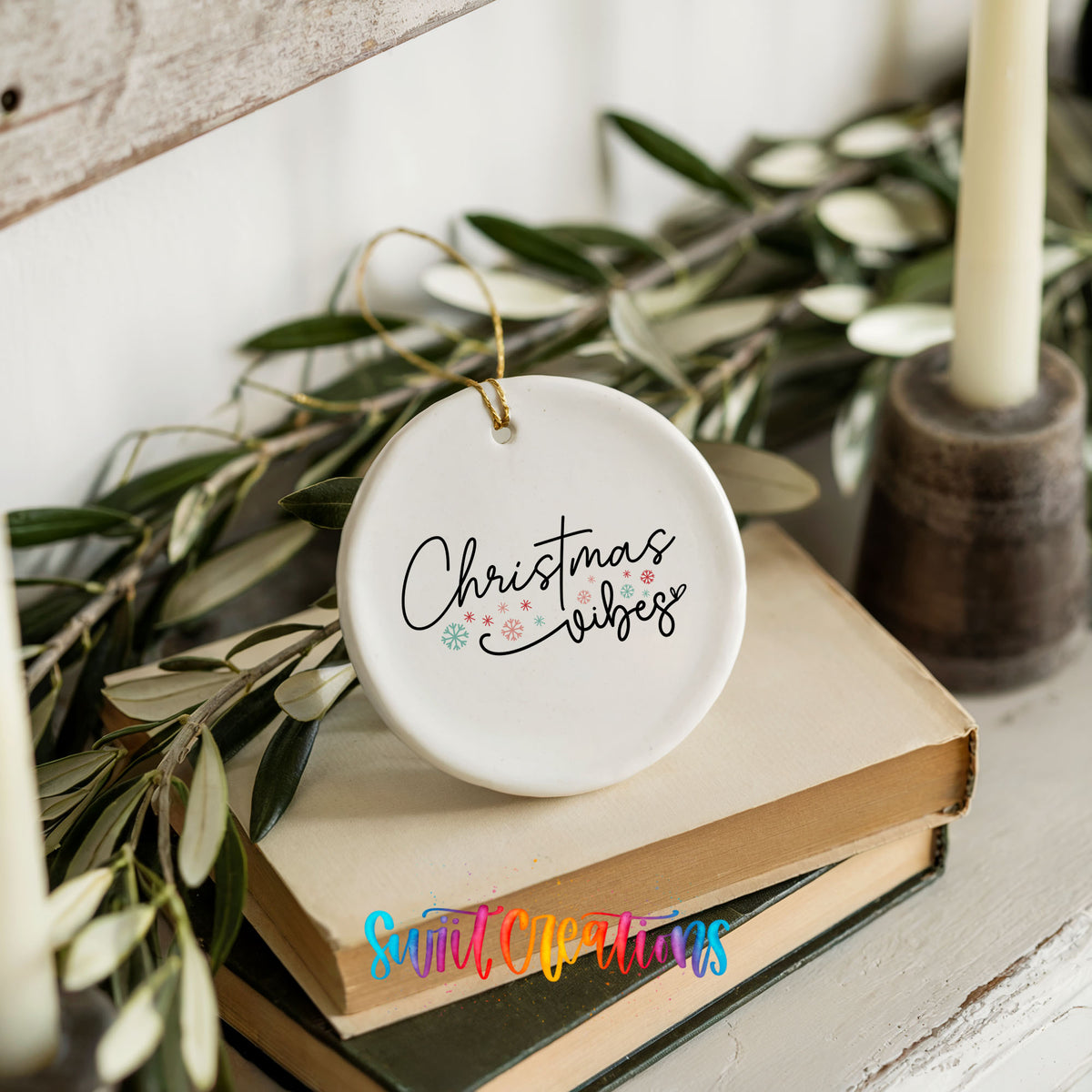 Round ceramic Christmas ornament featuring delicate Christmas Vibes script with small colorful holiday accents, styled on books with holiday greenery