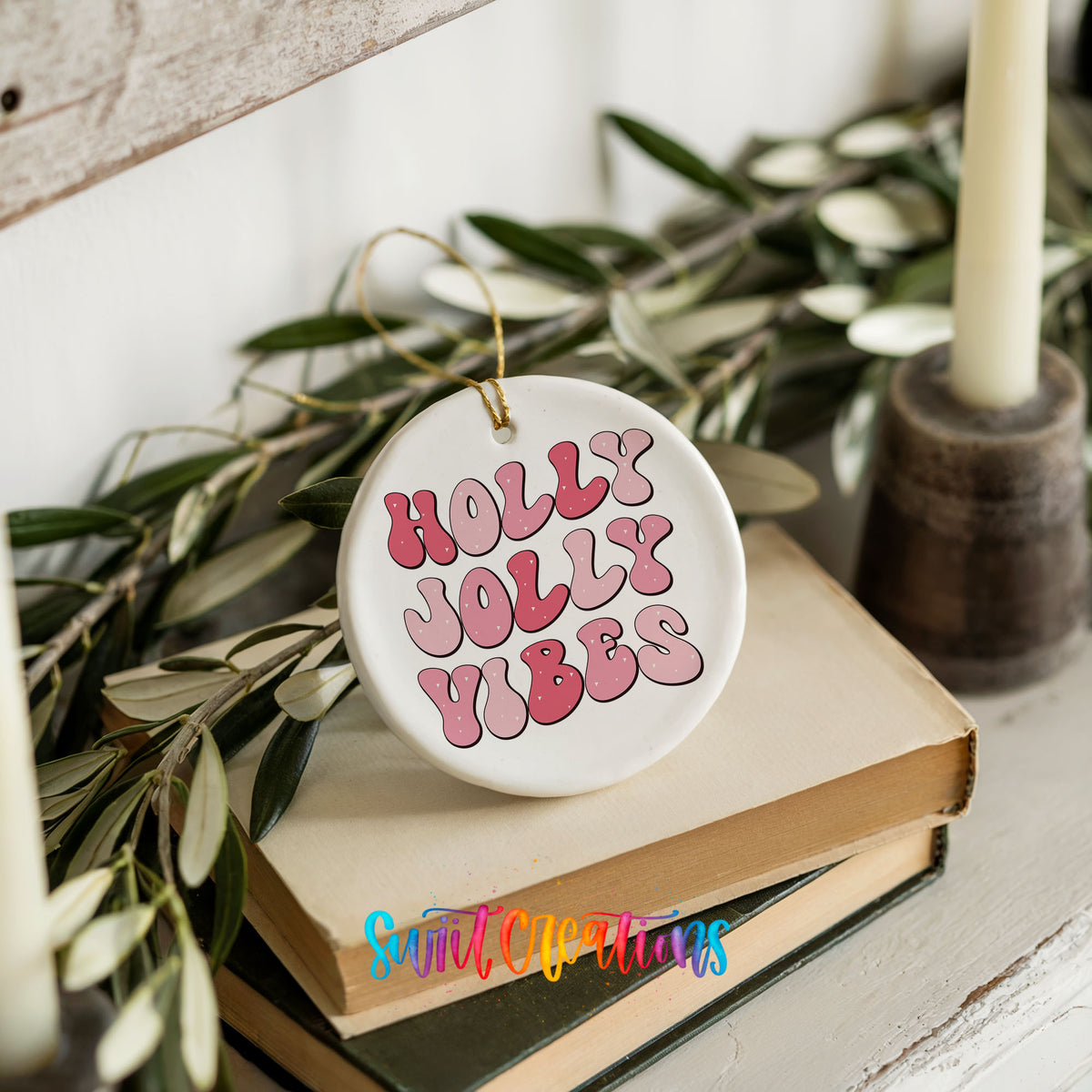 Round ceramic Christmas ornament featuring pink retro Holly Jolly Vibes typography, styled on books with holiday greenery