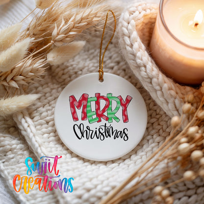 Round ceramic Christmas ornament featuring colorful patterned Merry Christmas typography, styled beside a candle and cozy knit blanket