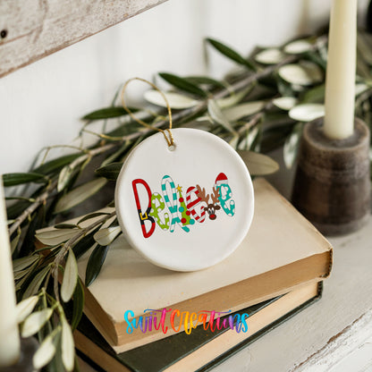 Round ceramic Christmas ornament featuring colorful whimsical Believe holiday lettering, styled on books with holiday greenery