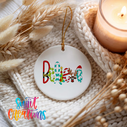 Round ceramic Christmas ornament featuring colorful whimsical Believe holiday lettering, styled beside a candle and cozy knit blanket