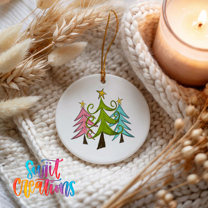 Round ceramic Christmas ornament featuring three colorful whimsical Christmas trees, styled beside a candle and cozy knit blanket