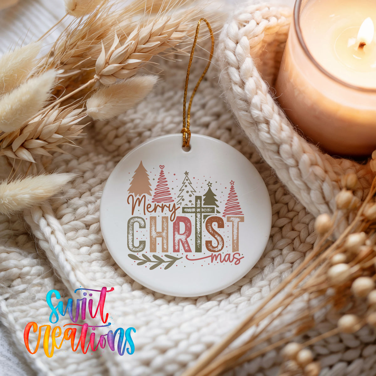 Round ceramic Christmas ornament featuring Merry Christmas typography with a pastel winter village and trees, styled beside a candle and cozy knit blanket