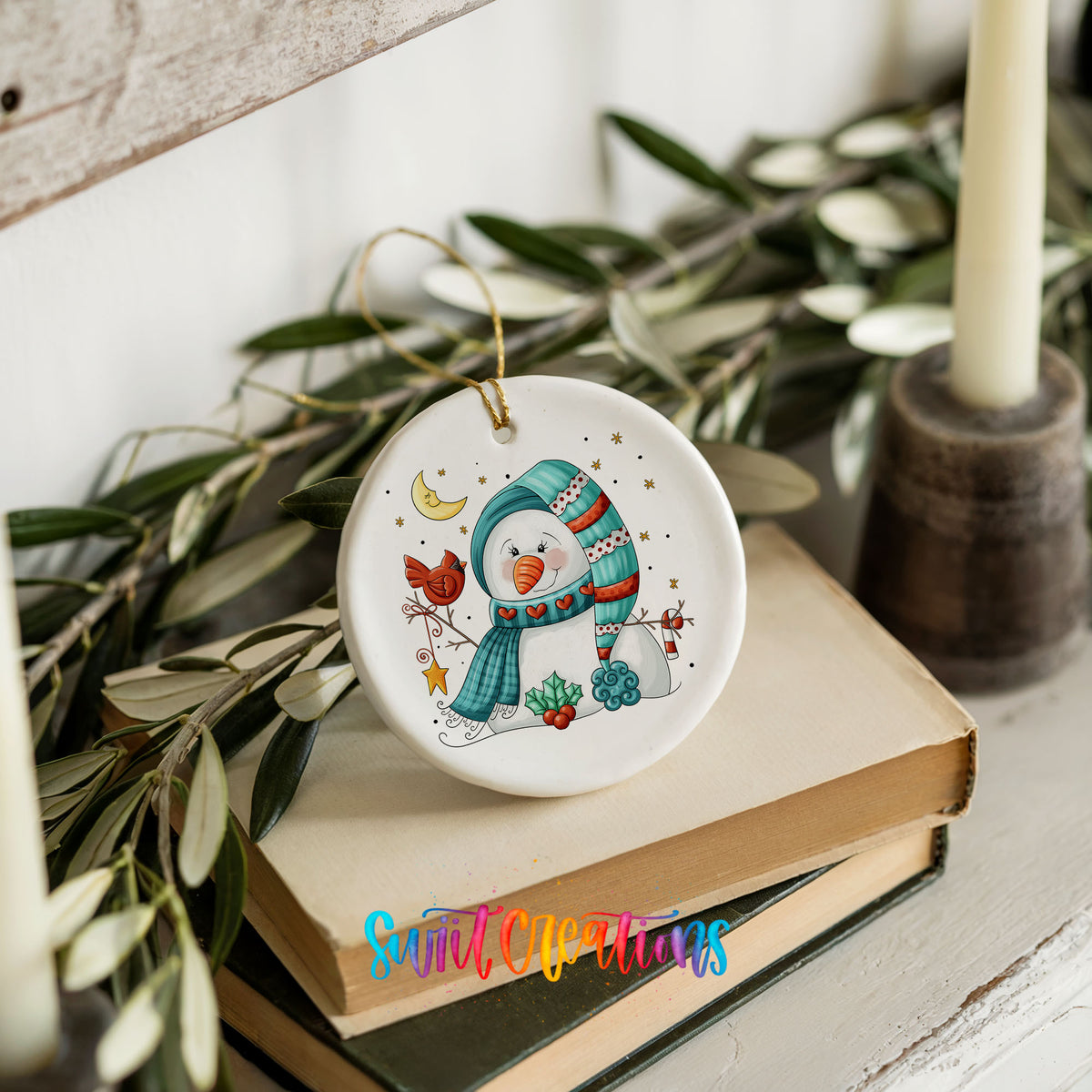 Round ceramic Christmas ornament featuring cozy snowman holding a bright red cardinal beneath a winter moon, styled on books with holiday greenery