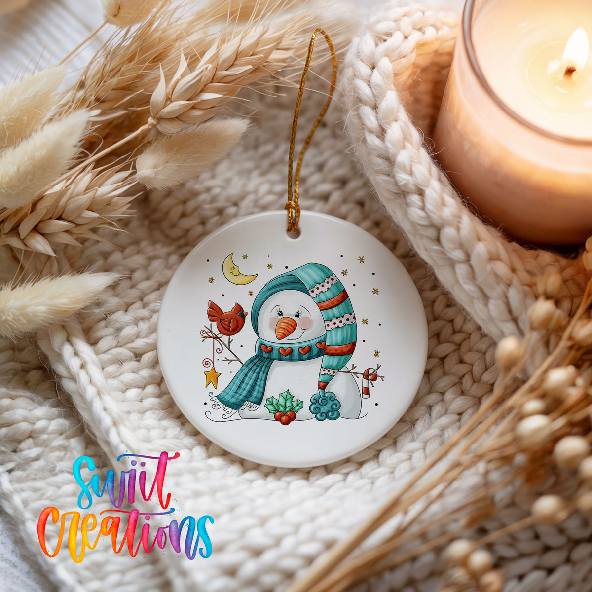 Round ceramic Christmas ornament featuring cozy snowman holding a bright red cardinal beneath a winter moon, styled beside a candle and cozy knit blanket
