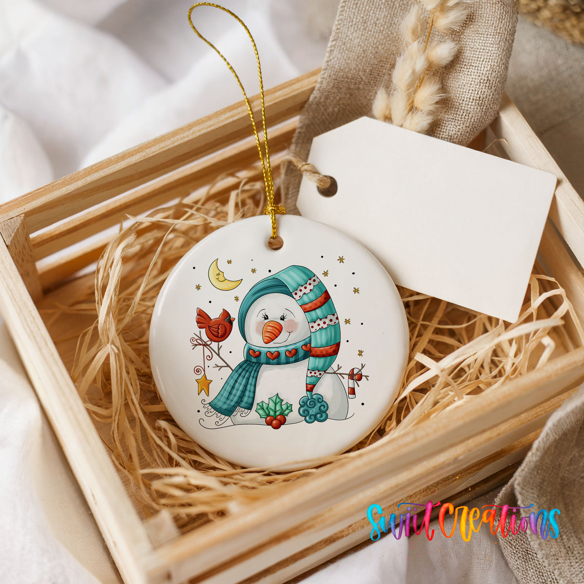 Round ceramic Christmas ornament featuring cozy snowman holding a bright red cardinal beneath a winter moon, presented in a rustic wooden gift crate