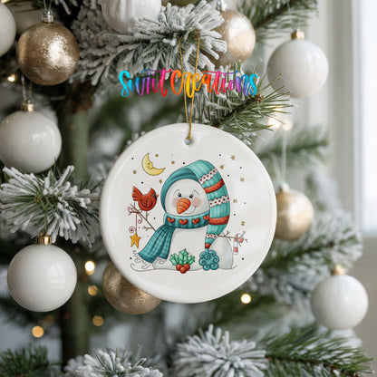 Round ceramic Christmas ornament featuring cozy snowman holding a bright red cardinal beneath a winter moon, hanging on a frosted holiday tree
