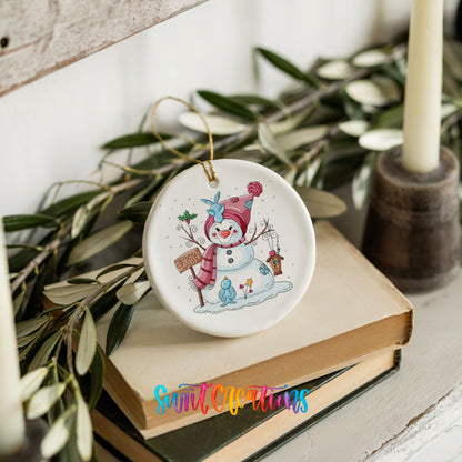 Round ceramic Christmas ornament featuring playful snowman dressed for a colorful winter snow day, styled on books with holiday greenery