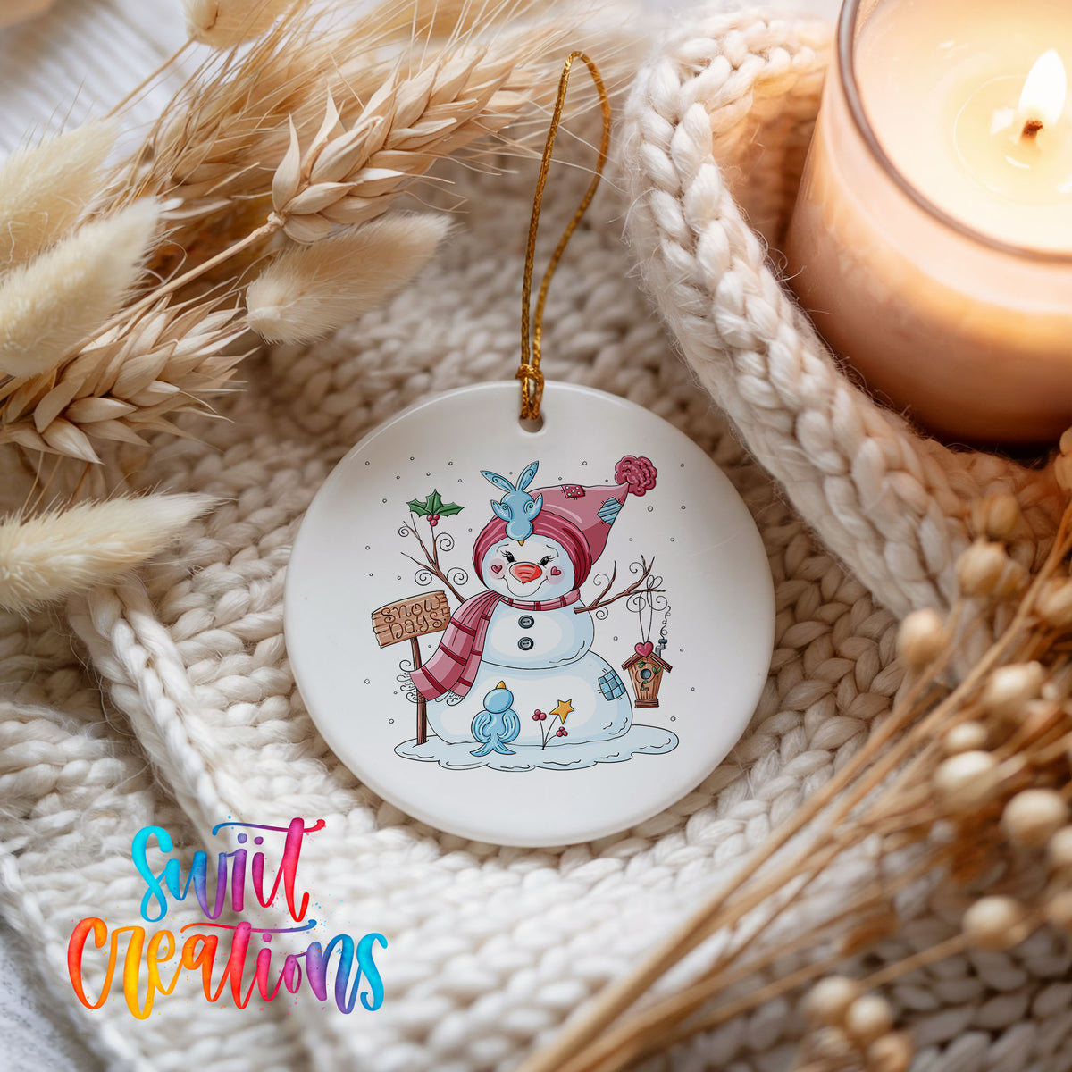 Round ceramic Christmas ornament featuring playful snowman dressed for a colorful winter snow day, styled beside a candle and cozy knit blanket