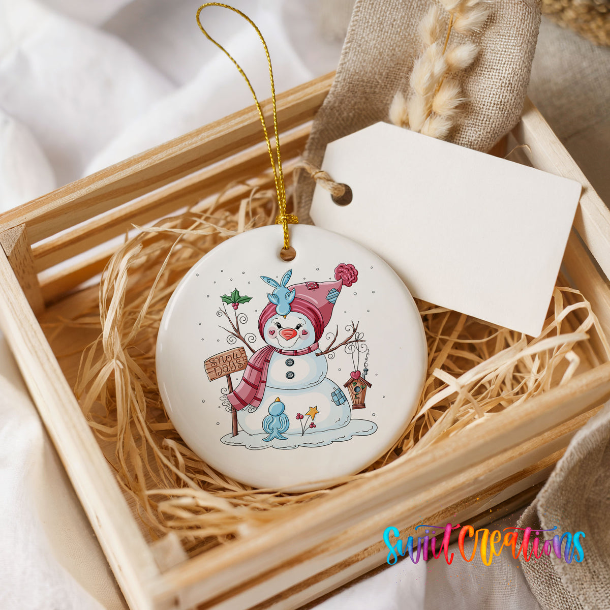 Round ceramic Christmas ornament featuring playful snowman dressed for a colorful winter snow day, presented in a rustic wooden gift crate