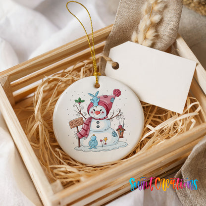 Round ceramic Christmas ornament featuring playful snowman dressed for a colorful winter snow day, presented in a rustic wooden gift crate