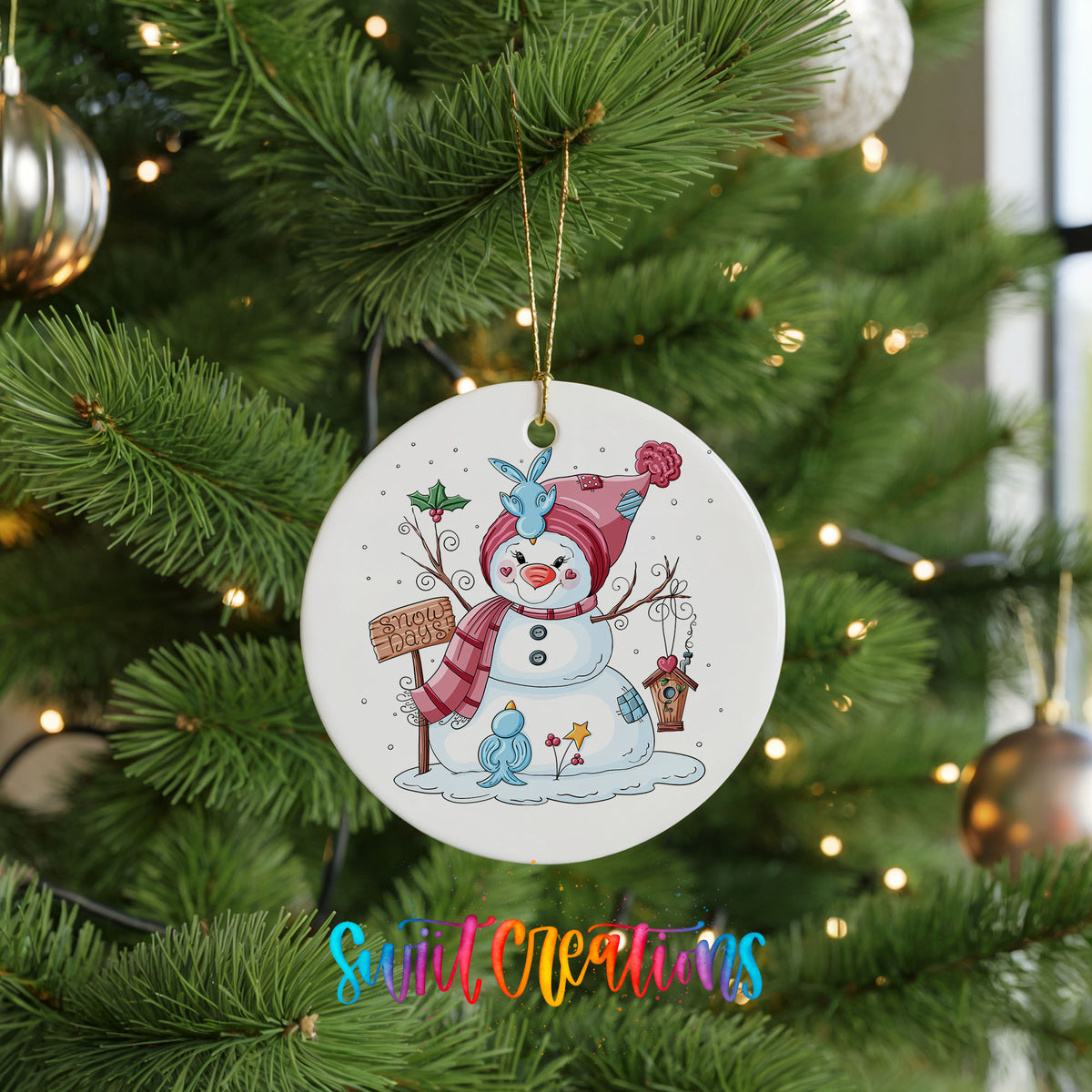 Round ceramic Christmas ornament featuring playful snowman dressed for a colorful winter snow day, hanging on a green Christmas tree