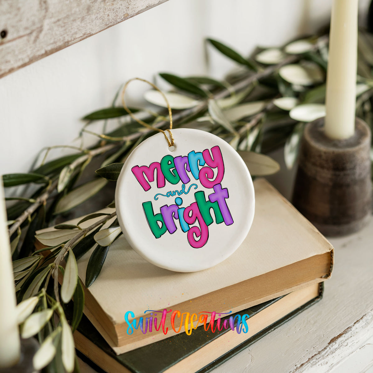 Round ceramic Christmas ornament featuring bright groovy Merry and Bright lettering in pink teal and purple, styled on books with holiday greenery